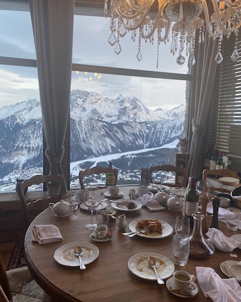 Una mesa de madera redonda con platos, copas, botellas y tetera. Detrás, una gran ventana ofrece vistas a montañas nevadas y un valle