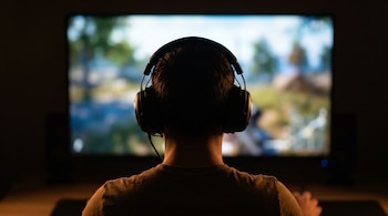 Vista trasera de una persona con auriculares, sentada en una habitación oscura mirando una pantalla grande con una imagen desenfocada de un paisaje natural.