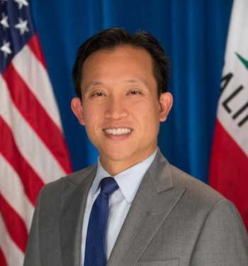 Retrato de David Chiu sonriendo con traje gris y corbata azul, con las banderas de Estados Unidos y California detrás