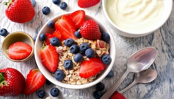 Tazón blanco con avena, arándanos y trozos de fresa, junto a un recipiente con yogur blanco, frutas dispersas y dos cucharas