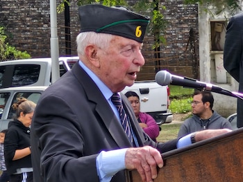 Primer plano de un hombre mayor con gorra militar y traje, hablando en un micrófono desde un podio de madera. Se ven otras personas y vehículos detrás