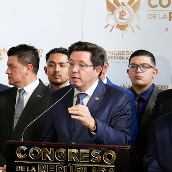 Hombre con traje azul oscuro, gafas, hablando en un micrófono desde un podio. Detrás, otros hombres en traje. Fondo blanco con escudo y texto "Congreso de la República"