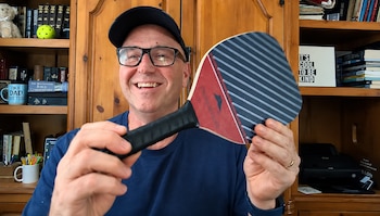 Un hombre con gafas y gorra negra sostiene una pala de pickleball de color rojo y azul frente a un mueble de madera con libros