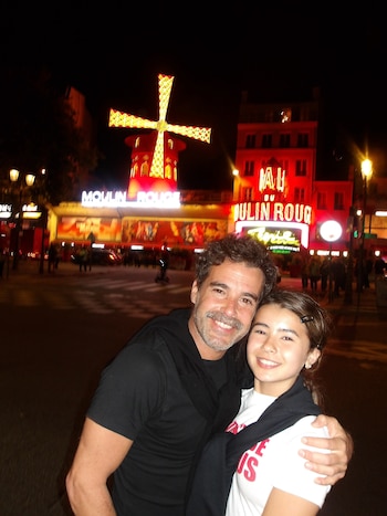 Un hombre y una joven abrazados y sonrientes posan en una calle nocturna delante del molino rojo e iluminado del Moulin Rouge
