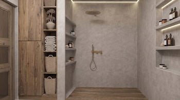 Ducha moderna con paredes de concreto gris, piso de madera, accesorios dorados. A la izquierda, un gabinete de madera con estantes, toallas apiladas y cestas.