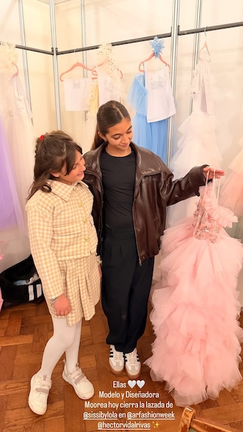 Dos niñas de pie frente a un perchero con ropa elegante; una viste traje a cuadros beige y la otra sostiene un largo vestido rosa de tul