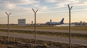 Una cerca de malla con un cartel de seguridad del aeropuerto y un avión comercial aterrizando al fondo.