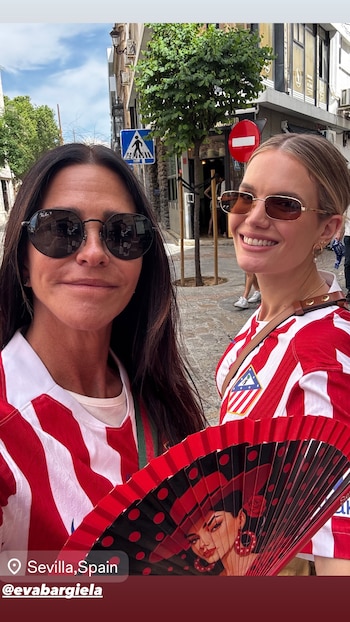 Carolina Baldini y Eva Barigela sonriendo, ambas con camisetas rojiblancas del Atlético de Madrid y gafas de sol, en una calle de Sevilla