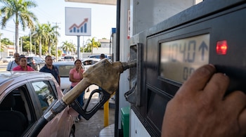 Primer plano de una mano operando un surtidor de gasolina que muestra un precio y una flecha ascendente, con un coche y personas al fondo en una estación.