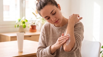 Una mujer de cabello recogido aplica crema blanca en su antebrazo izquierdo. Un tubo de crema se encuentra sobre una mesa de madera.