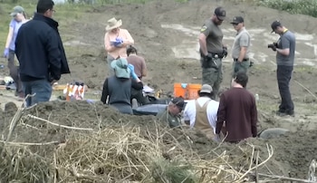 Grupo de personas en un área de tierra removida. Hay hombres uniformados, personas con guantes y otros tomando notas. Se ven herramientas, un cubo naranja y un objeto semienterrado