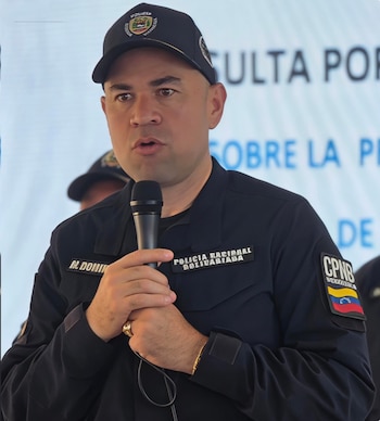 Primer plano de Miguel Domínguez, subdirector de la PNB, en uniforme oscuro, sosteniendo un micrófono. El fondo es una pantalla con texto borroso