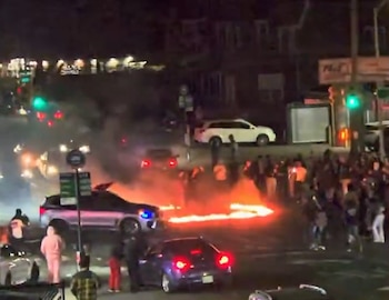 Vista nocturna de un coche gris realizando "donuts" con llamas rojas en el asfalto, rodeado de una multitud, otros vehículos y semáforos verdes