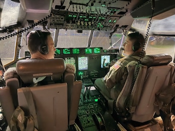 Parte frontal de un avión militar gris iluminado de noche en una pista de aterrizaje, con las escaleras de la puerta abiertas