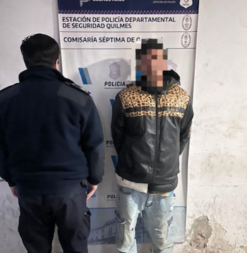 Un oficial de policía, de espaldas, junto a un hombre con una chaqueta negra y estampado de leopardo y jeans rotos, delante de un cartel de la Policía de Quilmes