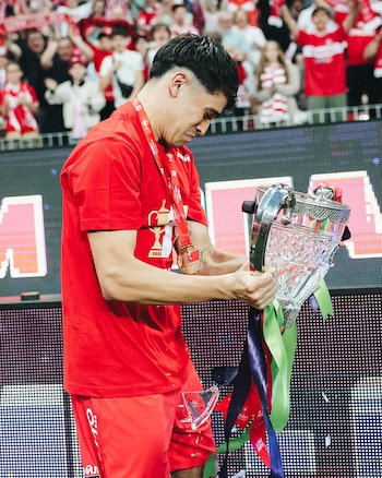 Un hombre con camiseta roja y medalla sostiene un trofeo de cristal roto. Fragmentos caen sobre sus shorts rojos, y se ven aficionados de fondo
