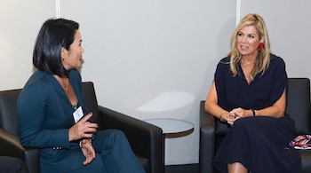 La reina Máxima de los Países Bajos y una mujer vestida con un traje oscuro conversan sentadas en sillones negros.