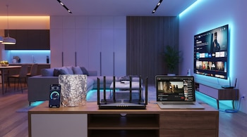 Un router WiFi, caja de papel aluminio, celular mostrando velocidad de internet, laptop y smart TV en un mueble dentro de una sala moderna con iluminación.