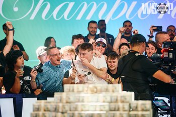 Un grupo de personas observa un evento. Un hombre se cubre la boca. Otros sostienen teléfonos y cámaras. Una pila de billetes de dólares en primer plano y un logotipo de WSOP en el fondo