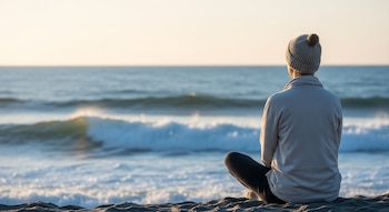 Vista trasera de una persona con gorro y suéter, sentada en la arena observando el mar con olas suaves y un cielo claro y luminoso.