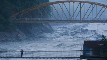 Un río de agua turbulenta fluye bajo un puente metálico amarillo y un puente colgante peatonal donde permanece una persona