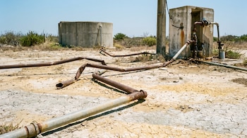 Tuberías de metal rotas y oxidadas sobre suelo seco y agrietado. Un tanque de agua de hormigón y conexiones deterioradas visibles al fondo, bajo un cielo azul.