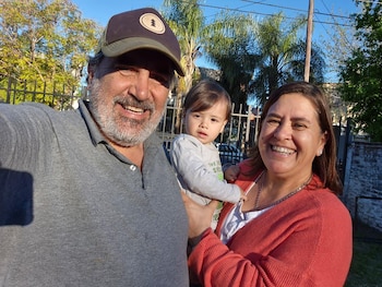 Hombre barbudo con gorra y mujer sonrientes sostienen a un bebé. Los tres miran a cámara y sonríen. Fondo borroso de árboles, un poste y una valla metálica