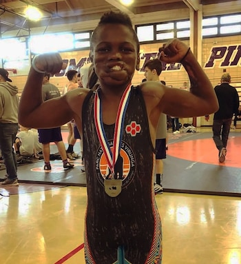 Un joven afroamericano en un gimnasio, con uniforme de lucha libre negro y detalles coloridos, flexiona sus bíceps y sonríe, llevando una medalla