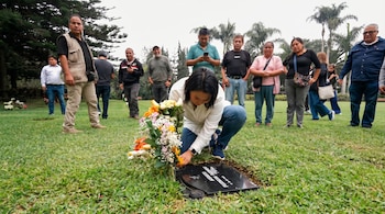Una mujer de camisa blanca y pantalón azul se agacha para colocar flores en una lápida negra. Personas observan alrededor. Césped, árboles y cielo claro.