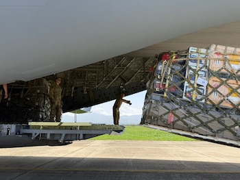 Avión de transporte con compuerta trasera abierta, tres soldados con uniforme militar, varios palés de carga con redes, pista de aterrizaje y montañas al fondo