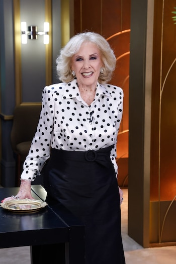 Mirtha Legrand sonríe, vestida con una blusa blanca de lunares negros y falda negra, posando con su mano sobre una bandeja dorada en un set de televisión