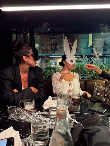 Dos mujeres sentadas a la mesa de un restaurante, una con un antifaz negro y otra con una máscara blanca de conejo