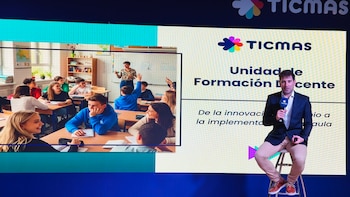 Hombre sentado en un taburete con micrófono frente a una pantalla. La pantalla muestra el logo TICMAS, texto sobre formación docente y una clase con alumnos