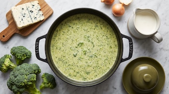 Olla negra con crema verde, ramitos de brócoli fresco, un trozo de queso azul, cebollas y una jarra sobre superficie de mármol.