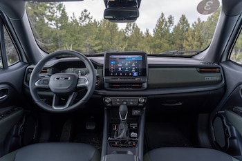 Interior de vehículo con volante, tablero digital, pantalla táctil, consola central, palanca de cambios, asientos negros y un bosque visible por el parabrisas