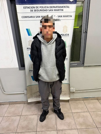 Hombre con rostro pixelado de pie frente a un banner de la Estación de Policía San Martín. Viste chaqueta negra, sudadera gris, pantalón gris y zapatillas negras