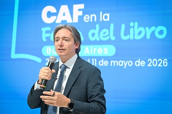 Primer plano de un hombre caucásico de mediana edad, con traje oscuro y corbata, hablando en un micrófono Shure. Detrás, una pantalla azul con texto blanco