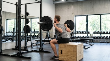 Hombre caucásico con camiseta gris y pantalones negros realiza sentadilla con barra, apoyando su trasero en un cajón de madera en un gimnasio.