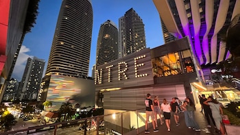 Vista aérea dentro de un centro comercial moderno de varios niveles con tiendas, escaleras mecánicas y personas caminando, con iluminación rosa y púrpura