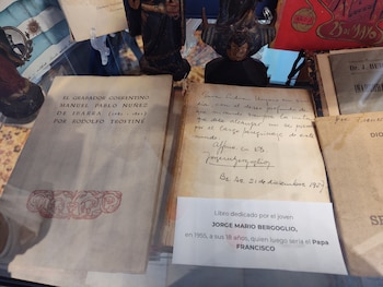 Dos libros antiguos expuestos en una vitrina junto a un letrero explicativo, una bandera argentina y estatuillas de madera