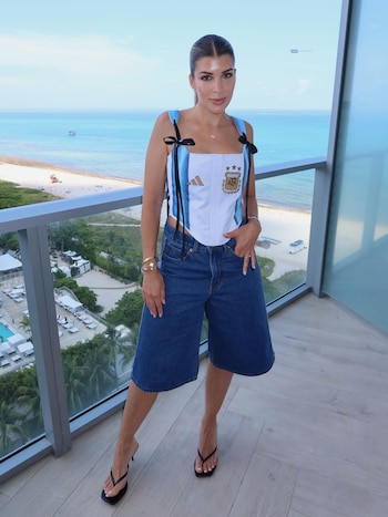 Mujer joven de pie en un balcón, con vista a la playa y el mar. Viste un top corset con el escudo de la selección argentina, pantalones de mezclilla y sandalias