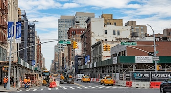 Vista de Flatbush Avenue con edificios, andamios, trabajadores de construcción, excavadoras y taxis. Señales del "Brooklyen Transformation Project" son visibles.