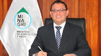 Hombre sonriente con traje de raya diplomática y corbata a rayas azules y blancas, sentado en una silla, con un bolígrafo y una bandera del MINAGRI