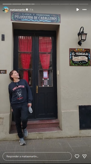Matías Martín, vestido con una sudadera oscura, se apoya en una pared beige frente a la entrada de una barbería con puerta negra y cortinas rojas