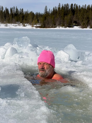 Wim Hof, un hombre barbudo con gorro rosa, inmerso hasta el pecho en agua helada con bloques de hielo flotando. Al fondo, un bosque nevado bajo cielo claro