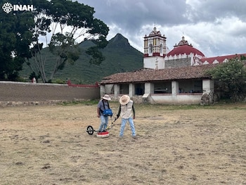 Dos personas con sombrero operan equipo de estudio geofísico en un campo. Al fondo, edificio histórico colonial con cúpula roja y montañas