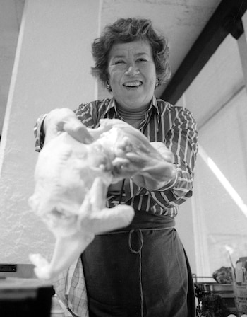 Fotografía en blanco y negro de una mujer sonriente con pelo rizado, camisa de rayas y delantal que sujeta un pollo crudo
