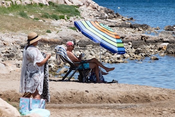 Un hombre disfruta de la playa bajo su sombrilla mientras una mujer a su lado prepara la toalla para bañarse en el mar.