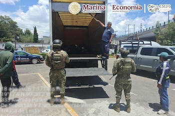 Dos marinos uniformados observan un camión de carga abierto junto a vehículos oficiales en una calle soleada