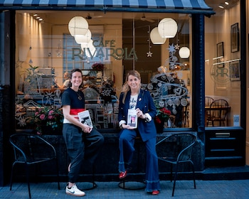 Dos mujeres sentadas frente a la vidriera de una cafetería con sillas de metal sostienen un libro cada una